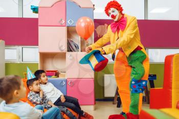Funny clown shows tricks with balloon to surprised children. Birthday party celebrating in playroom, baby holiday in playground. Childhood happiness, childish leisure