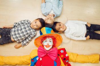 Funny clown and little children lying on the floor and pull their hands into the camera, top view. Birthday celebrating, baby holiday in playground. Childhood happiness