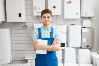 Plumber in uniform at showcase with boilers in plumbering store. Man with notebook buying sanitary engineering in shop, water taps and faucets choice