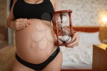 Pregnant woman with belly poses against sandglass at home, side view. Pregnancy, calm in prenatal period. Expectant mom resting in bedroom, healthy life