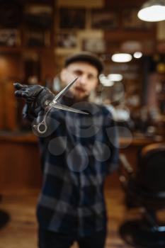 Barber in hat and gloves shows sciccors. Professional barbershop is a trendy occupation. Male hairdresser in retro style hairdressing salon