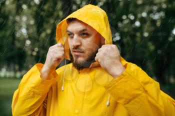 Man in raincoat puts on a hood, summer park in rainy day. Alone male person in rain cape on walking path, wet weather in alley