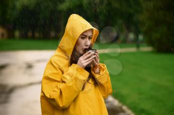 Woman in raincoat drinks hot tea in summer park, rainy day. Alone female person in rain cape on walking path, wet weather in alley