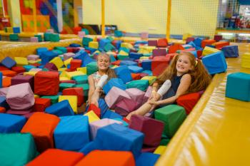 Two little girls lying in soft cubes, playground in entertainment center. Play area indoors, playroom