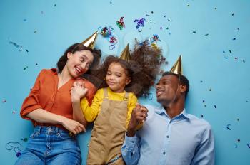 Funny family in caps celebrate birthday, blue background. Pretty little girl and her parents, event celebration, balloons and confetti decoration