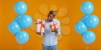 Cheerful man in cap holding two gift boxes with red ribbons, yellow background. Smiling male person got a surprise, event or birthday celebration, balloons decoration