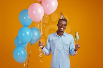Funny man in cap holding a glass of beverage, yellow background. Smiling male person got a surprise, event or birthday celebration, balloons decoration