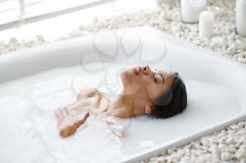 Attractive woman relax, skincare in bath with milk. Female person in bathtub, beauty and health care in spa, wellness treathment in bathroom, pebbles and candles on background