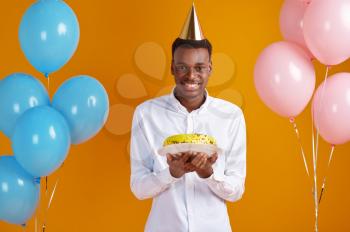 Cheerful man in cap with birthday cake, yellow background. Smiling male person got a surprise, event celebration, balloons decoration