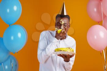 Cheerful man in cap with smeared face tasting birthday cake, yellow background. Smiling male person got a surprise, event celebration, balloons decoration