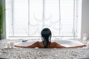 Attractive young woman relax in a bubble bath, back view. Female person in bathtub, beauty and health care in spa, wellness treathment in bathroom, pebbles and candles on background
