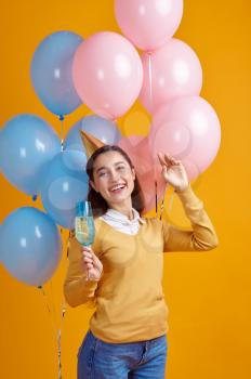 Funny woman in cap holding a glass of beverage, yellow background. Pretty female person got a surprise, event or birthday celebration, balloons decoration