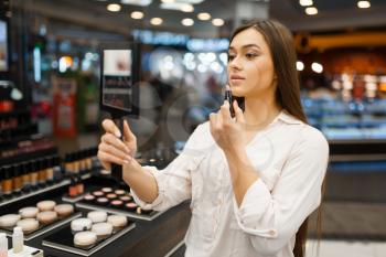 Attractive woman with mirror applies lipstick in cosmetics store. Buyer at the showcase in luxury beauty shop salon, female customer in fashion market