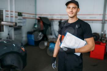 Auto repairman in uniform holds inspection report, tire service. Technician repairs car tyre in garage, professional automobile inspection in workshop, vehicle on lift jack on background