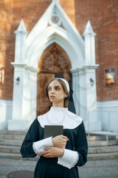 Young nun in a cassock holds a book, church on background. The sister is preparing for prayer in the monastery, religion and faith, religious person