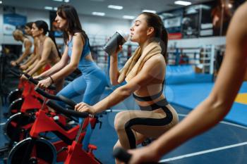 Woman drinks water on a stationary bike in gym. People on fitness workout in sport club, athletic girls in sportswear on training indoors