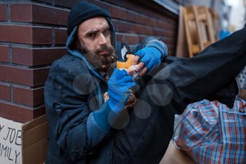 Bearded beggar eats burger on city street. Poverty is a social problem, homelessness and loneliness, alcoholism and drunk addiction, urban lonely