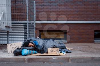 Bearded dirty homeless with help sign lies on city street. Poverty is a social problem, homelessness and loneliness, alcoholism and drunk addiction, urban lonely