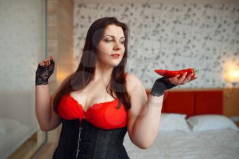 Fat perverse woman in erotic lingerie holds red pepper. Sexy overweight girl with big breast, corrupt large size lady