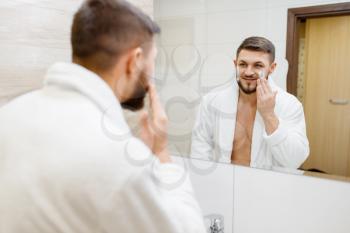 Man in bathrobe applies shaving foam at the mirror in bathroom, routine morning hygiene. Male person at the sink performs skin and body treatment procedures