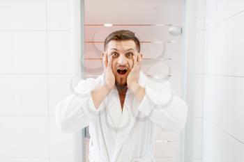 Bearded man frightened by his appearance at the mirror in bathroom, morning hygiene. Male person at the sink performs skin and body treatment procedures