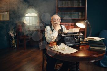 Elderly writer works on vintage typewriter in his home office. Old man in glasses writes literature novel in room with smoke