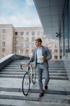 Businessman in suit with bicycle comes down the stairs at the office building in downtown. Business person riding on eco transport on city street, urban style