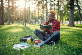 Black student in glasses reading book on the grass in summer park. A teenager studying outdoors and having lunch