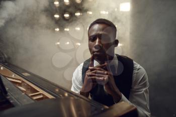 Ebony grand piano musician poses on the stage with spotlights on background. Negro pianist poses at musical instrument before concert