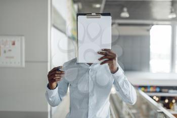 Ebony businessman covers his face with a notebook in mall. Successful business person, black man in formal wear, shopping center