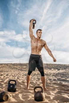 Muscular male athlete doing exercises with kettlebell in desert at sunny day. Strong motivation in sport, strength outdoor training, sportsman with weights