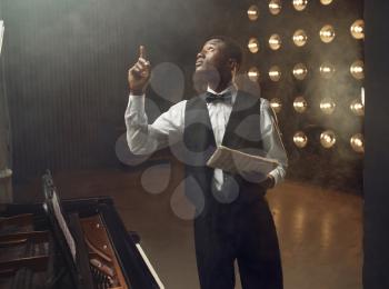 Ebony grand piano player with music notebook in his hands on the stage with spotlights on background. Negro performer poses at musical instrument before concert