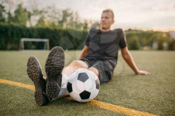 Male soccer player with ball sitting on the grass on the field. Footballer on outdoor stadium, workout before game, football training