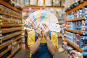 Male builder showing color palette in hardware store. Constructor in uniform look at the goods in diy shop, paint department