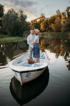 Love couple standing in a boat on quiet lake at summer day on sunset. Romantic meeting, boating trip, man and woman walking along the lake