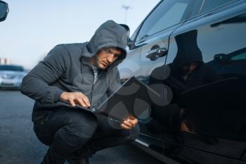 Car thief with laptop hacking alarm system, criminal lifestyle. Hooded male robber opening vehicle on parking. Auto robbery, automobile crime