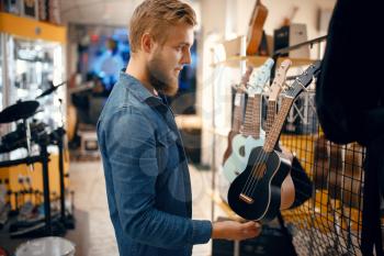 Bearded young man choosing ukulele guitar in music store. Assortment in musical instruments shop, male musician buying equipment
