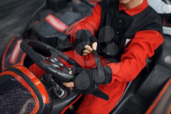 Kart racer in red uniform puts on gloves, karting auto sport indoor. Speed race on close go-kart track with tire barrier. Fast vehicle competition, high adrenaline hobby