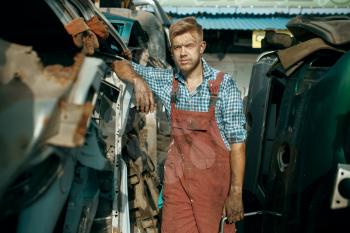 Male repairman poses on car junkyard. Auto scrap, vehicle junk, automobile garbage, abandoned, damaged and crushed transport