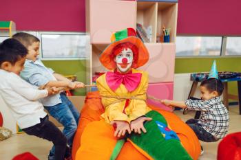 Joyful little boys tied up funny clown in children's area. Birthday party celebrating in playroom, baby holiday in playground. Childhood happiness, childish leisure