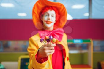 Funny clown with lollipops in hand poses in children's area. Birthday party celebrating in playroom, baby holiday in playground. Childhood happiness, childish leisure, entertainment with animator