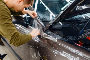 Male worker installs transparent protection film on car hood. Installation of coating that protects the paint of automobile from scratches. New vehicle in garage, tuning