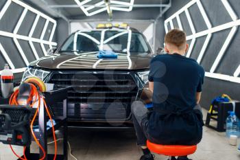 Male worker polishes car surface using polishing machine, detailing. Preparationa before installation of coating that protects the paint of automobile from scratches. Vehicle in garage, auto tuning