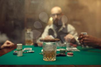 Whiskey and cigar on gaming table with green cloth, bearded poker player on background, casino. Games of chance. Man leisures in gambling house