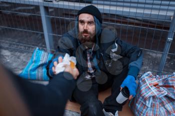 Male person gives food to bearded dirty beggar on city street. Poverty is a social problem, homelessness and loneliness, alcoholism and drunk addiction, urban lonely