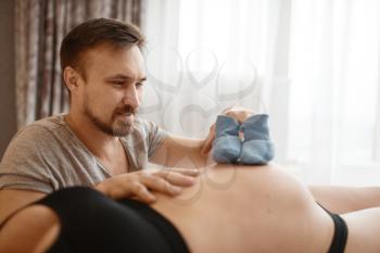 Happy couple, pregnant wife and husband plays with clothing for newborns on belly at home, bedroom interior on background. Pregnancy, prenatal period. Expectant mom and dad are resting in bed