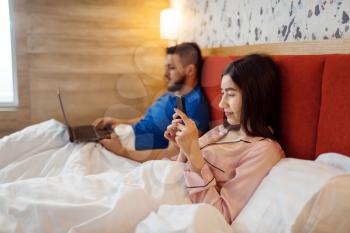 Love couple in pajamas lying in bed and using their gadgets, good morning. Harmonious relationship in young family. Man and woman resting together in their house, carefree weekend