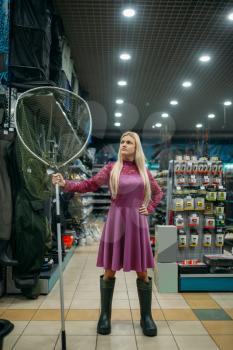 Fisherwoman in rubber boots holds net in fishing shop, hooks and baubles on background. Equipment and tools for fish catching and hunting, accessory choice on showcase in store, bait assortment