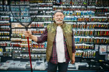 Happy angler holds net in fishing shop, hooks and baubles on background. Equipment and tools for fish catching and hunting, accessory choice on showcase in store, bait assortment