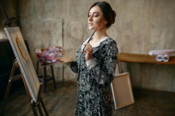 Female painter with brush and palette poses in art studio. Artist at her workplace, creative master at the easel in workshop